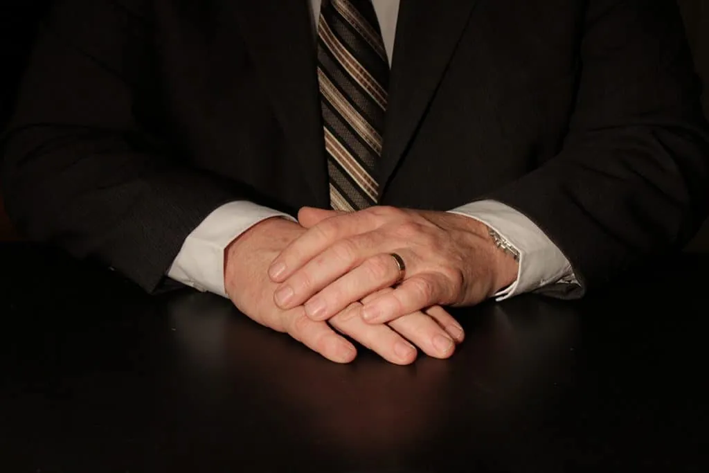 Man wearing a dark suit and striped tie with hands clasped on a black table