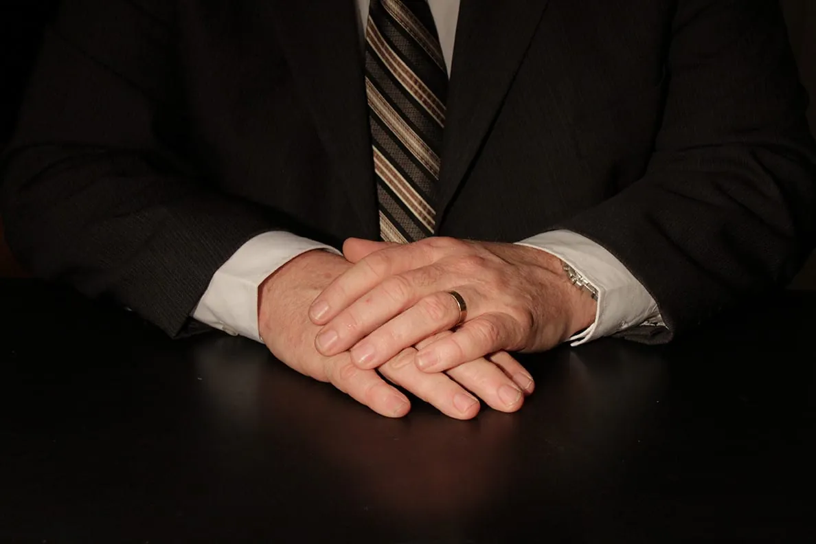 The Importance of Hands in Communication (Part One) 3 Crossed hands of a man wearing a black suit and a striped tie, on a dark table