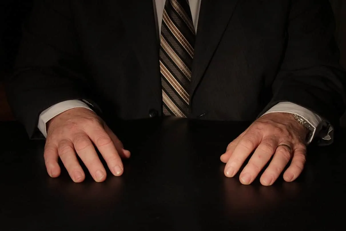 The Importance of Hands in Communication (Part Two) 2 A man in a suit with his hands resting on a black table, wearing a watch and a striped tie