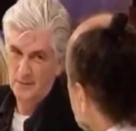 White-haired man looking at another person, seen from behind, during a conversation